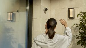 Een vrouw bij een voordeur zwaait naar een Hue Secure beveiligingscamera die aan de muur is bevestigd. 