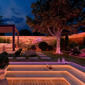 En hage og uteplass opplyst med myke toner av gult, rosa og lilla lys fra skjulte lysremser under trappetrinn og synlige stripeys langs blomsterbed og på vegger. 