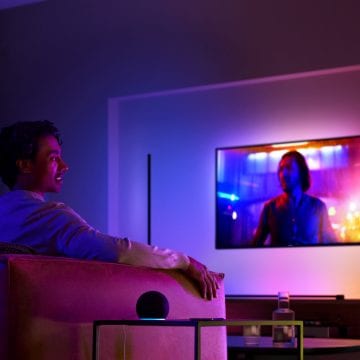 Mężczyzna na kanapie oglądający telewizję w pomieszczeniu z oświetleniem przestrzennym i inteligentnym głośnikiem stojącym na stoliku obok